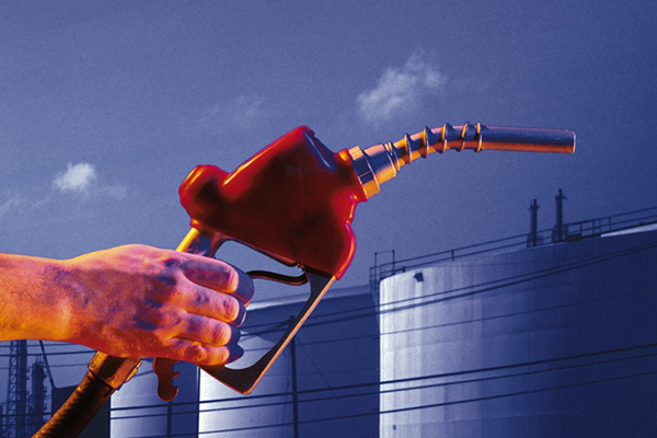 A hand holding a red gas pump handle with oil storage containers in the background