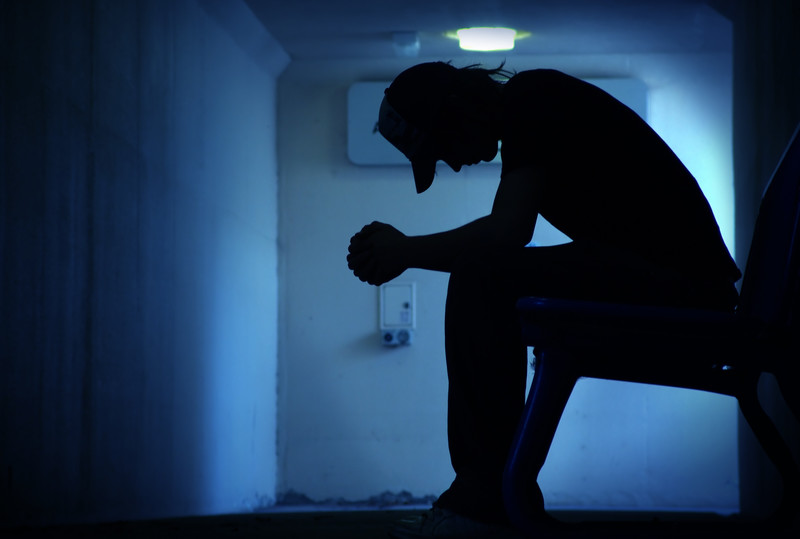 Silhouette of man in ball cap sitting in small dimly lit room head bowed and hands clasped