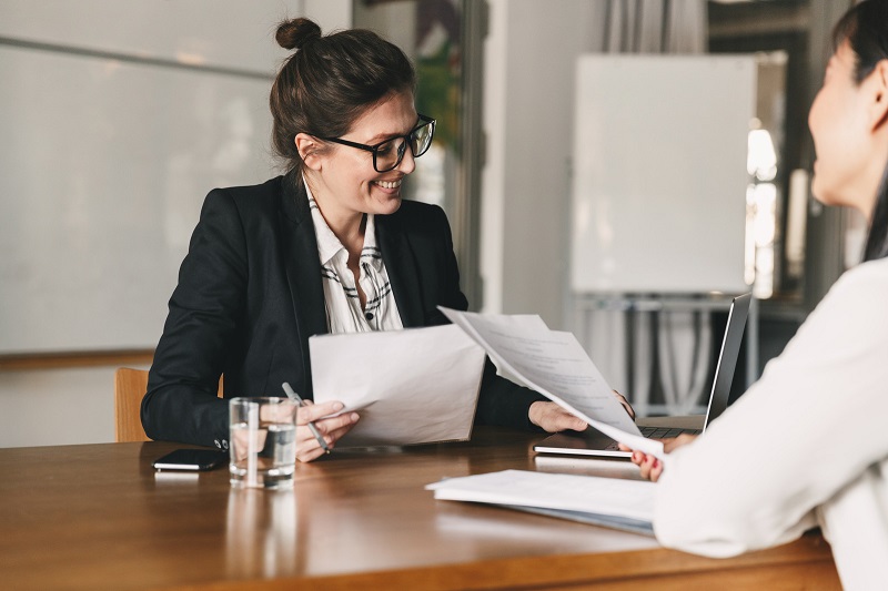 Woman sitting at desk holding papers talking to second woman