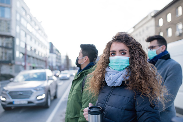 Three people standing on the sidewalk of a busy street wearing medical masks