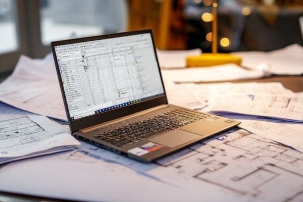 laptop with spreadsheets on the screen surrounded by construction blueprints