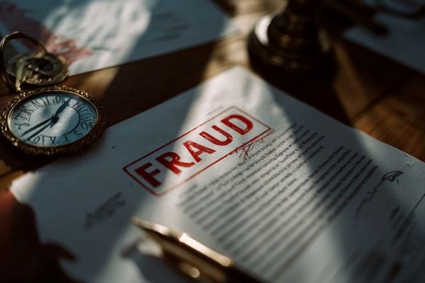 official document with "FRAUD" stamped in red next to a timepiece and a pen on a wooden desk