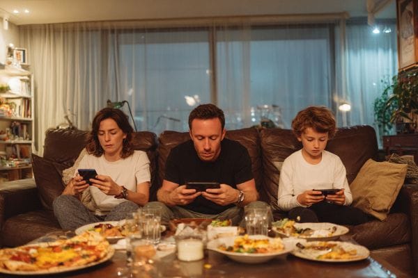 three family members in their living room sitting on a couch with food staring at their smartphones