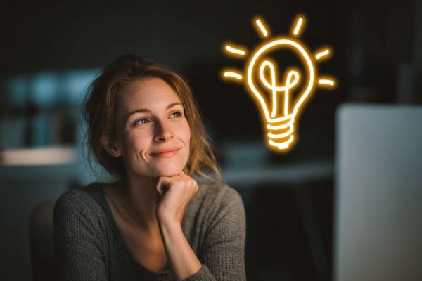 woman sitting at her desk looking up at a floating neon lightbulb drawn in the air